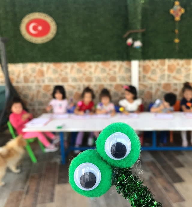 Pompon gözlü el işi kuklası, arkada masada çalışan çocuklar